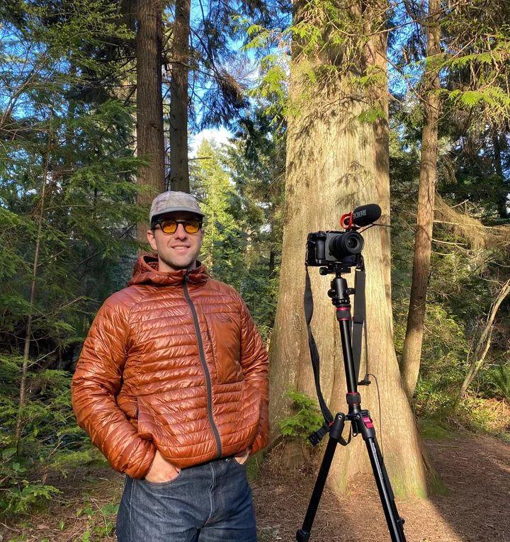 Graeme in an orange puffer jacket beside a camera on a tripod, among tall trees in a forest